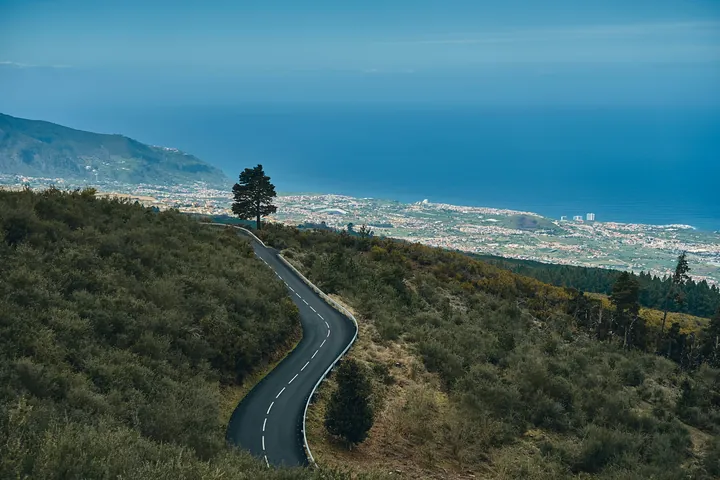 A winding asphalt road on a mountain slope overlooking a town on Tenerife and the Atlantic Ocean. The hillside is forested, the sky and the sea are blue.	